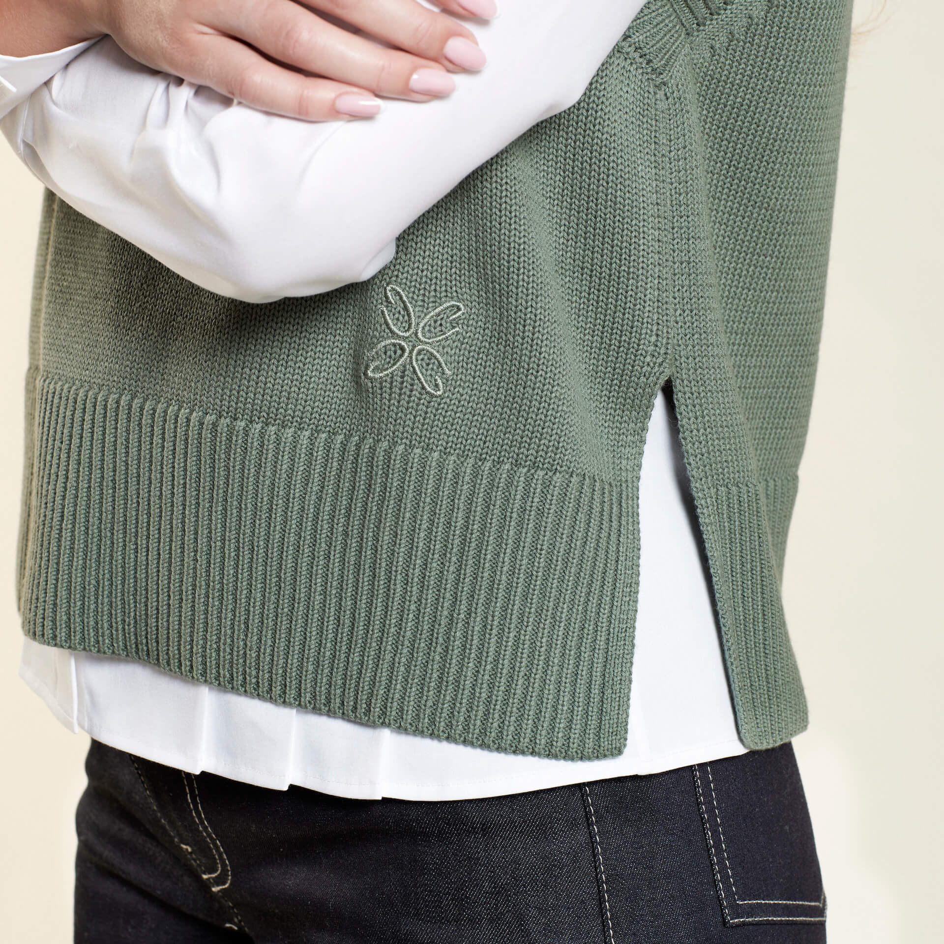 Close-up of a green knit vest from CYME Copenhagen, featuring a side slit and subtle embroidered logo on the lower side, layered over a white shirt and dark denim jeans. The image highlights the intricate details and quality of the garment.