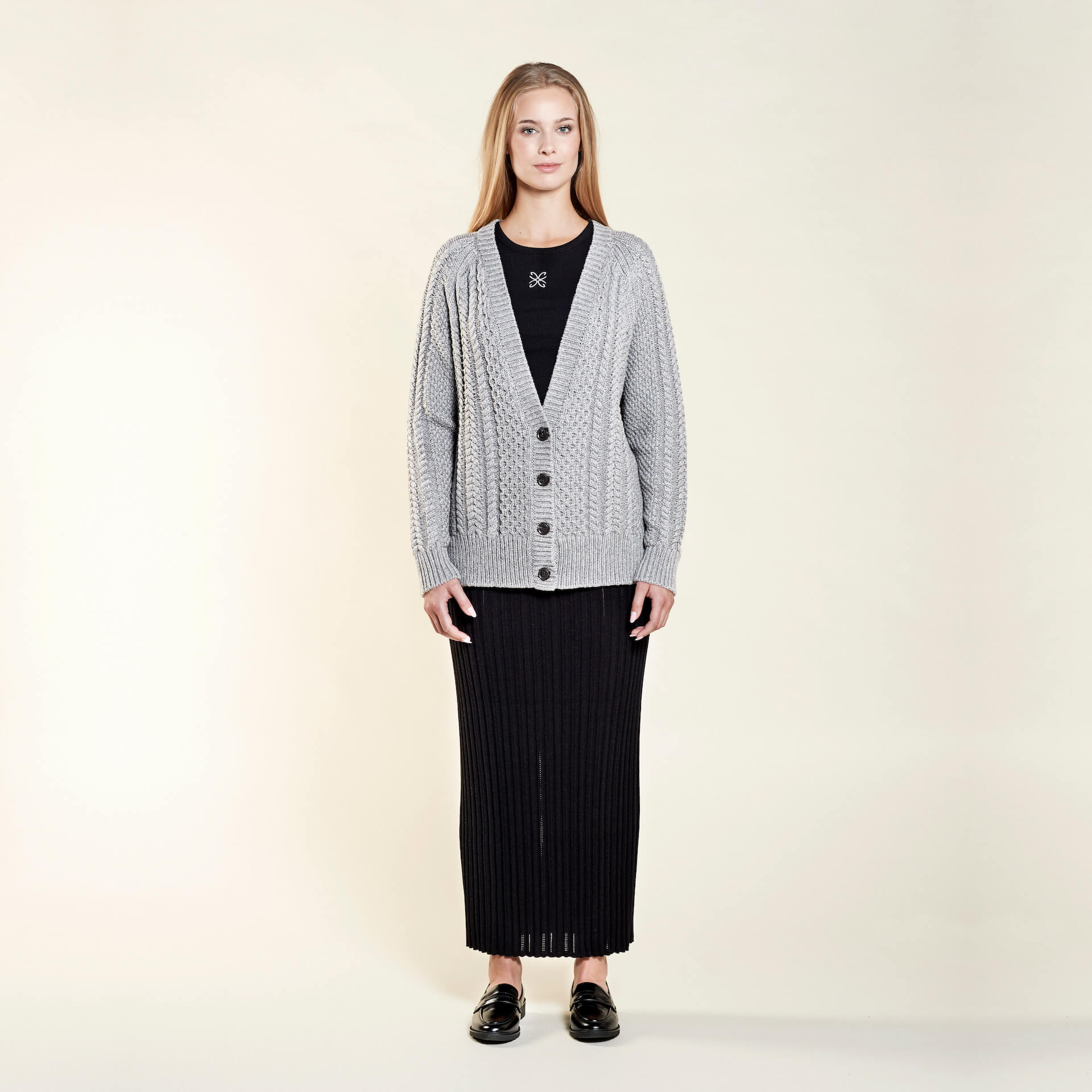 A woman with long blonde hair models a cozy outfit from CYME Copenhagen. She is wearing a light grey cable-knit cardigan over a black ribbed dress, paired with black loafers. The ensemble highlights the brand's focus on comfort and timeless style.