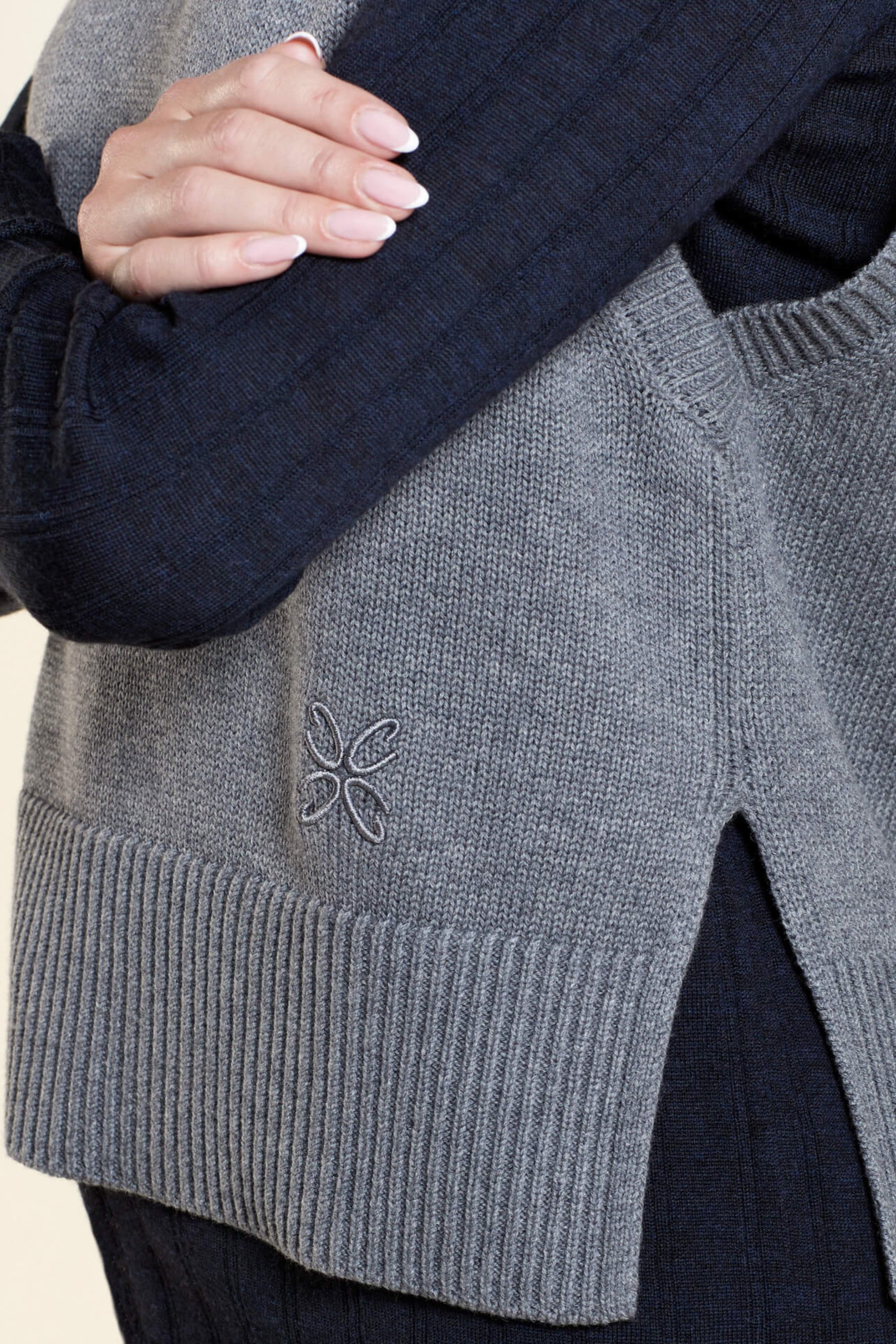 Close-up of a grey knit vest from CYME Copenhagen, featuring a side slit and subtle embroidered logo on the lower side, layered over a dark long-sleeve shirt. The image highlights the detailed craftsmanship and quality of the knitwear.