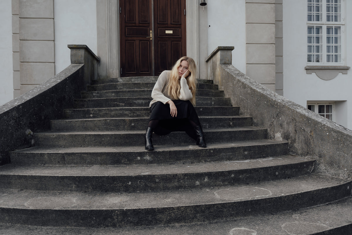 Model wearing a light knit jumper and black skirt, seated on stone steps