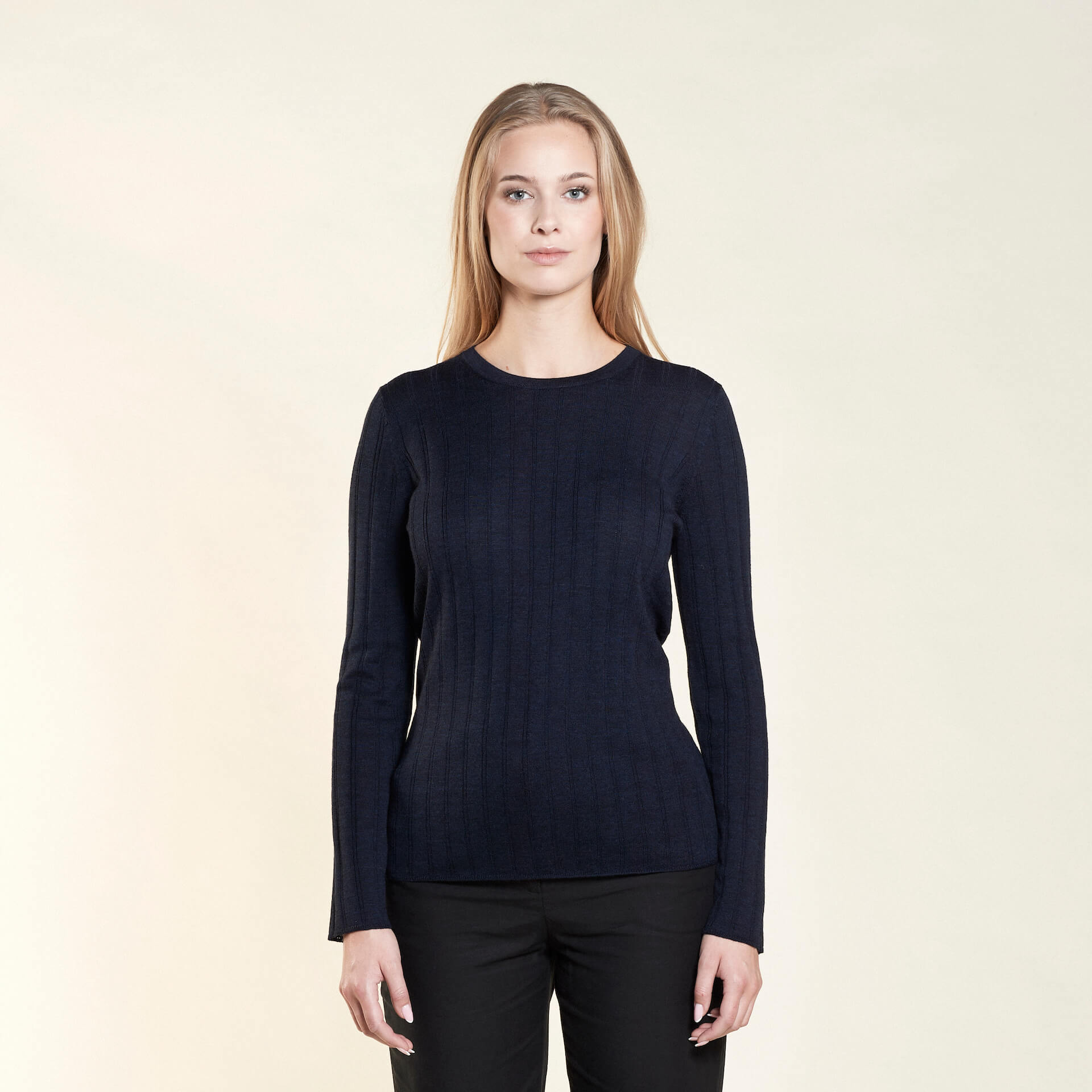 A woman with long blonde hair models a simple and elegant outfit from CYME Copenhagen. She is wearing a navy blue ribbed long-sleeve sweater paired with black trousers, emphasizing the brand's focus on timeless and high-quality fashion.