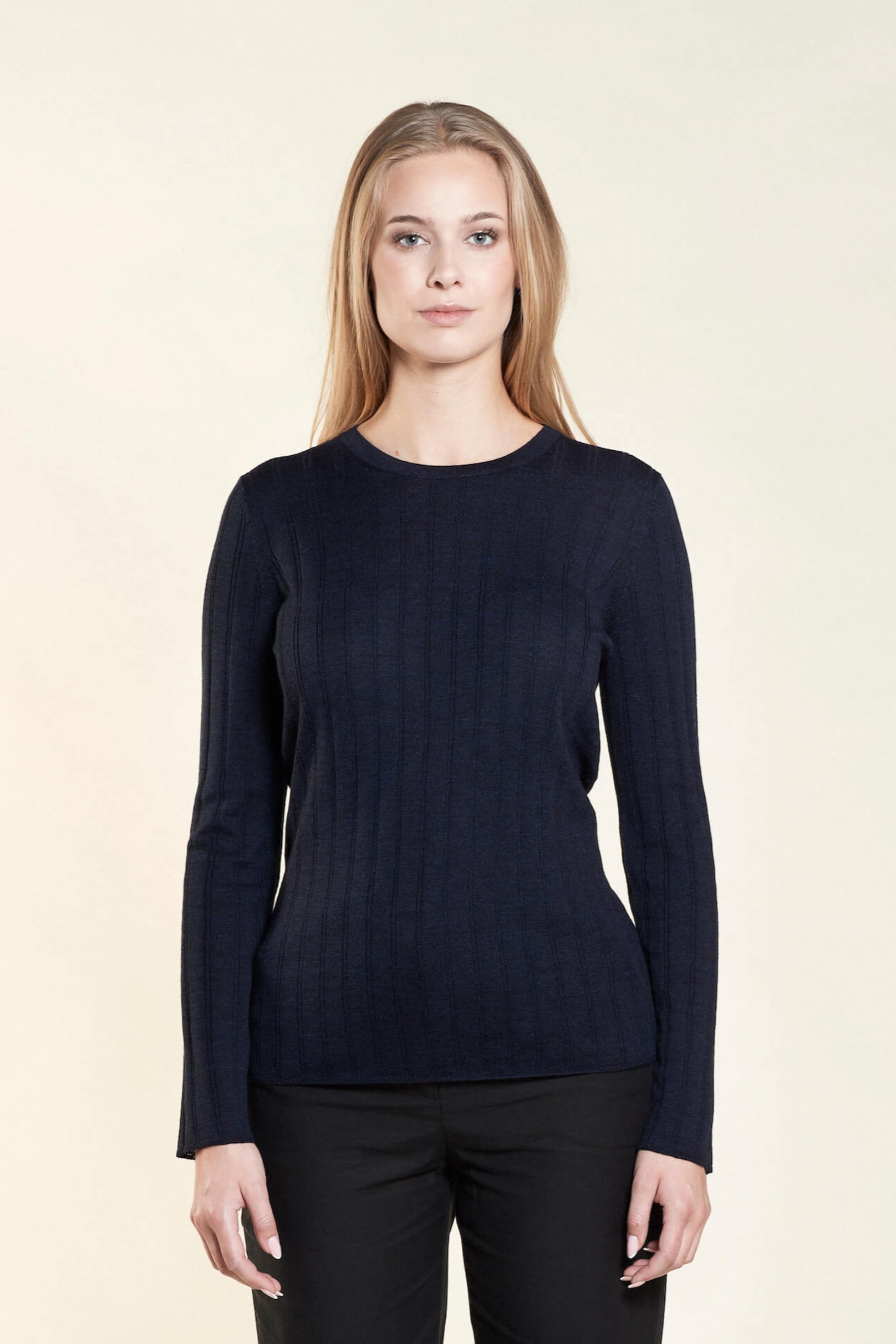 A woman with long blonde hair models a simple and elegant outfit from CYME Copenhagen. She is wearing a navy blue ribbed long-sleeve sweater paired with black trousers, emphasizing the brand's focus on timeless and high-quality fashion.