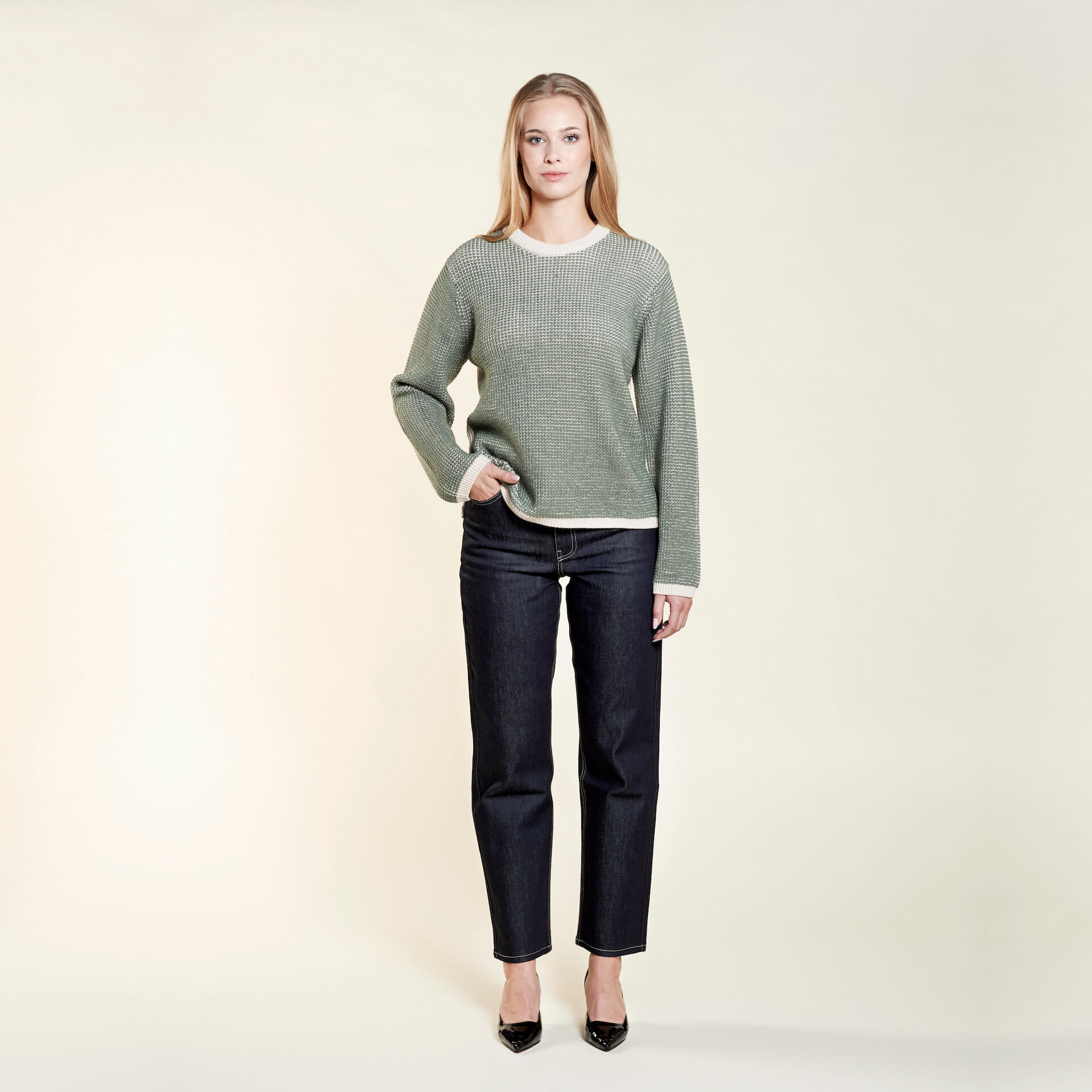 A woman with long blonde hair models a casual outfit from CYME Copenhagen. She is wearing a sage green textured long-sleeve sweater with contrasting white trim, paired with dark blue jeans and black heels. The outfit highlights the brand's focus on timeless and comfortable fashion.