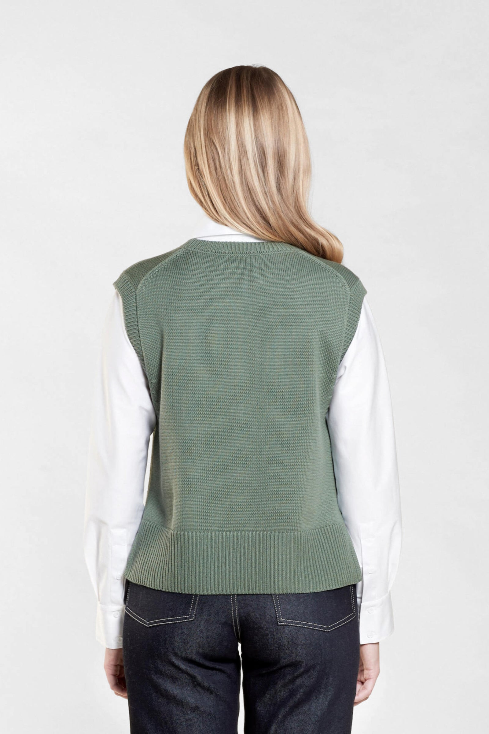 Rear view of a woman with long blonde hair modeling a sage green sleeveless knit vest over a white collared shirt, paired with dark blue jeans from CYME Copenhagen. The image highlights the vest's relaxed fit and the clean lines of the outfit, emphasizing the brand's focus on timeless, versatile fashion.