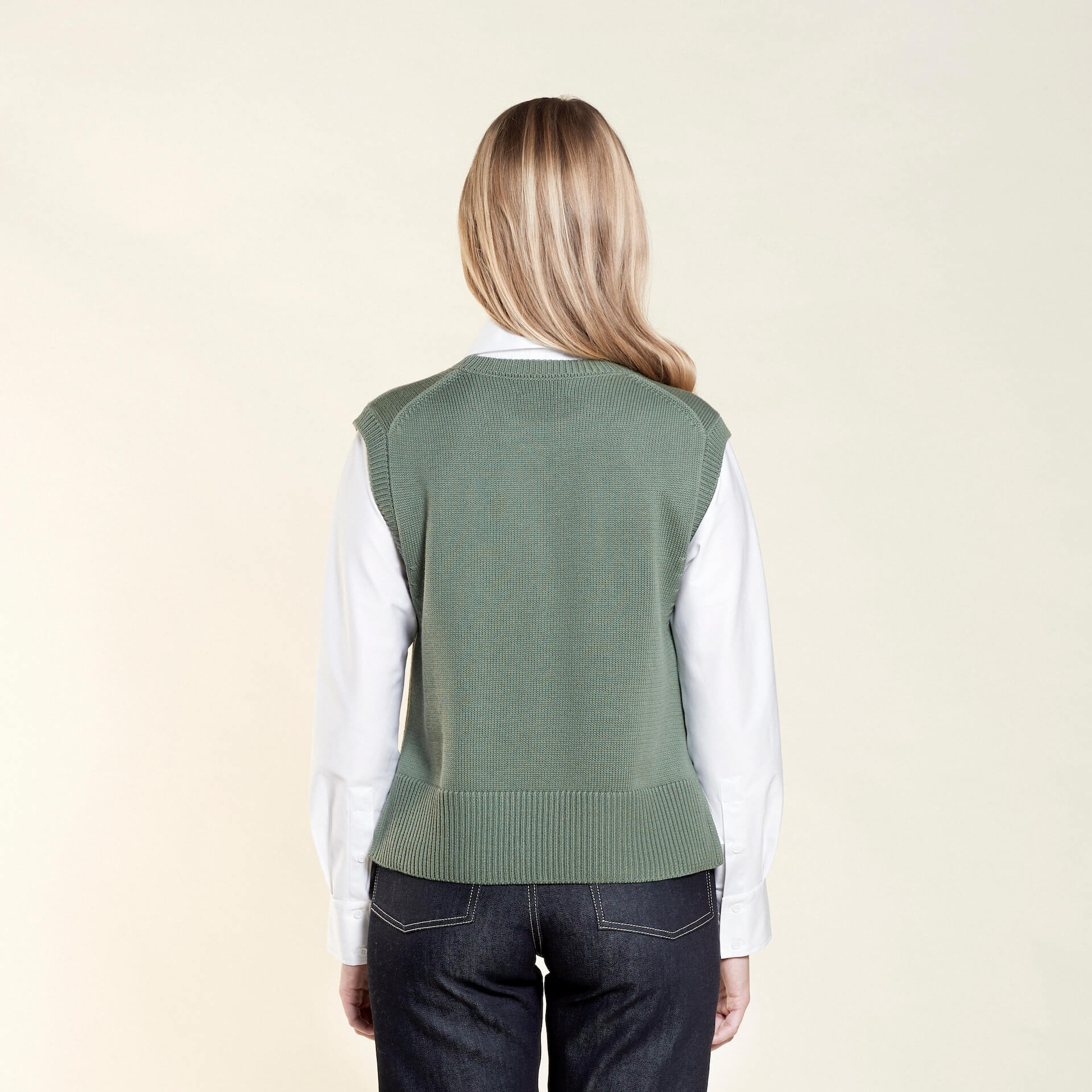 Rear view of a woman with long blonde hair modeling a sage green sleeveless knit vest over a white collared shirt, paired with dark blue jeans from CYME Copenhagen. The image highlights the vest's relaxed fit and the clean lines of the outfit, emphasizing the brand's focus on timeless, versatile fashion.