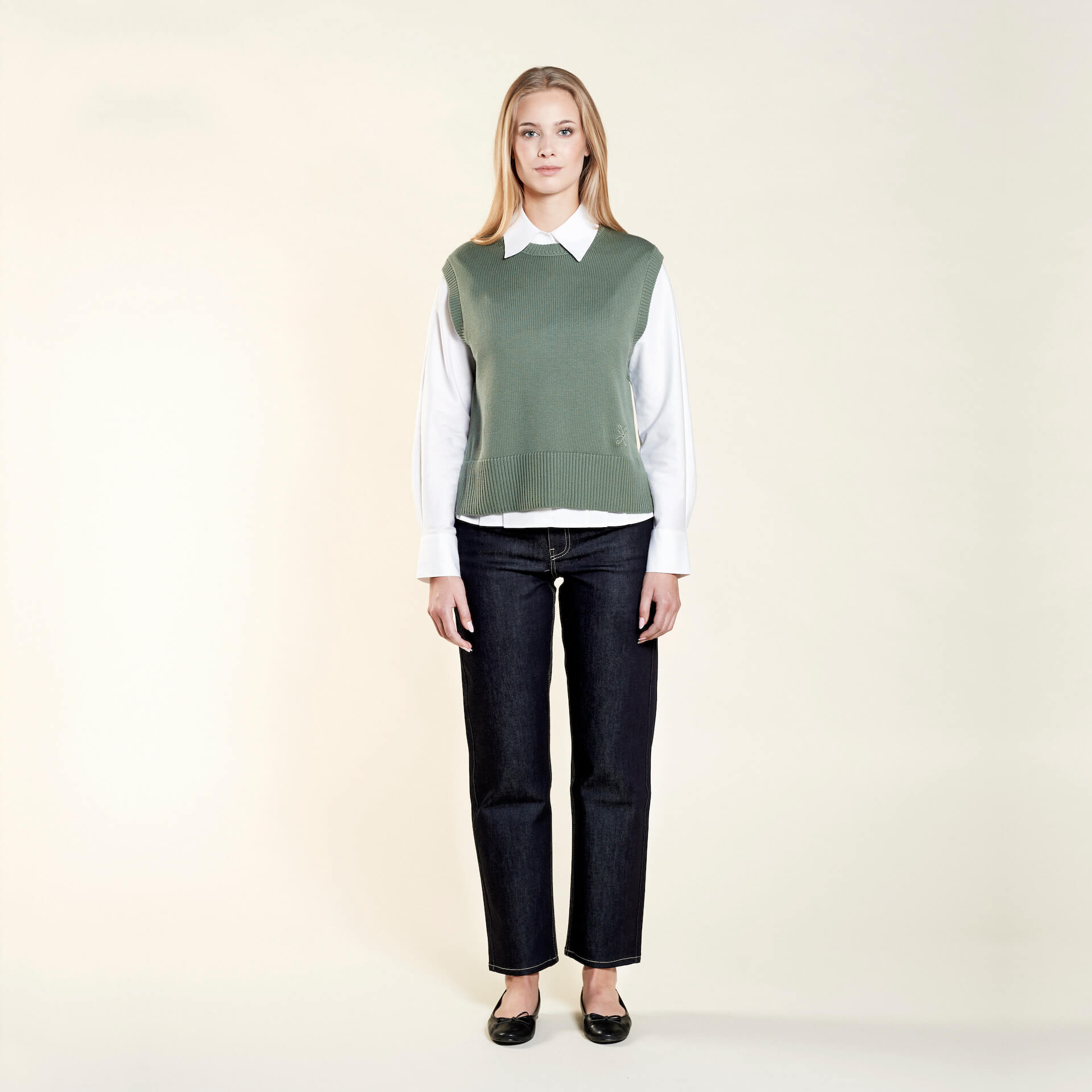 A woman with long blonde hair models a stylish outfit from CYME Copenhagen. She is wearing a sage green sleeveless knit vest over a white collared shirt, paired with dark blue jeans and black ballet flats. The outfit highlights the brand's focus on timeless and versatile fashion.