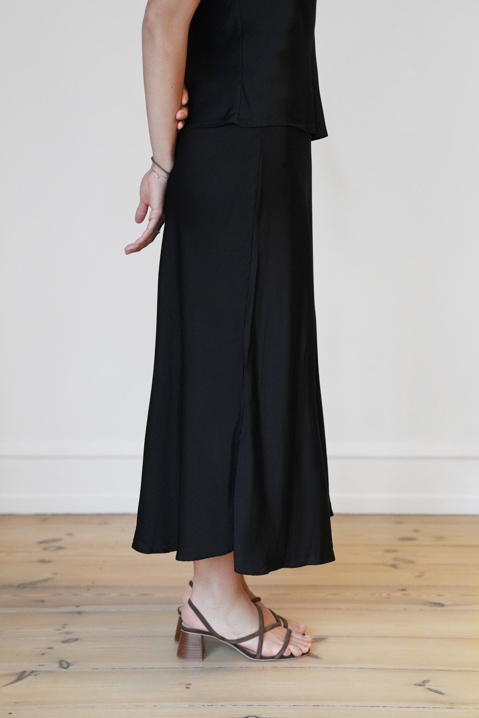 Side view of the CYME Satin Skirt in black. A high-waisted, ankle-length skirt with a fluid drape and subtle flare—made from elegant satin for refined movement and shine.