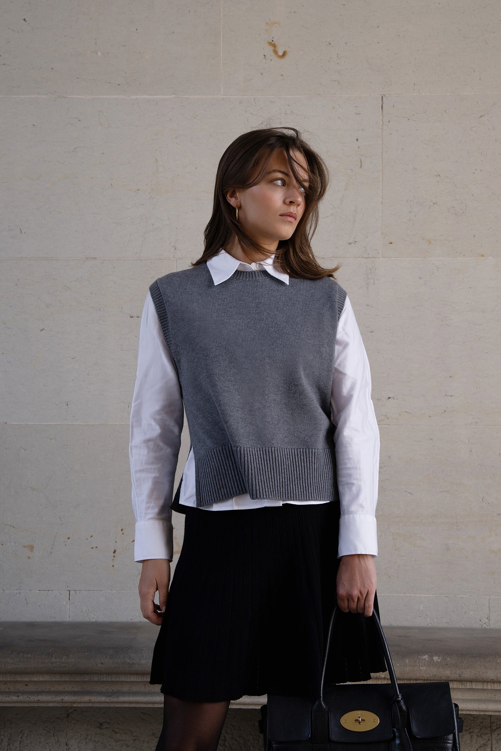 Woman wearing a gray sleeveless sweater over a white shirt with a black skirt, standing against a plain wall.