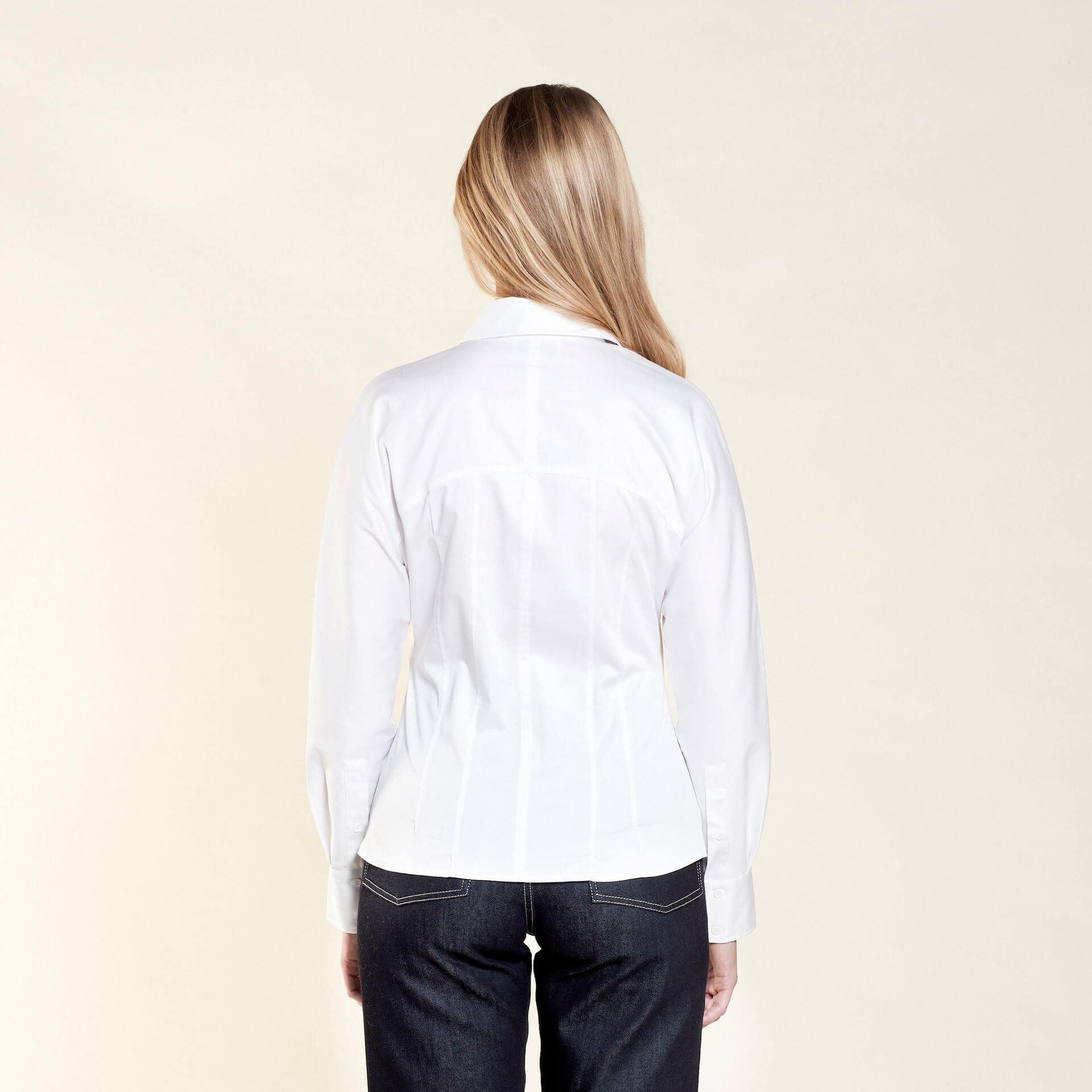 Rear view of a woman with long blonde hair modeling a white button-down shirt paired with dark blue jeans from CYME Copenhagen. The image highlights the shirt's tailored fit and clean lines, emphasizing the brand's focus on high-quality craftsmanship.