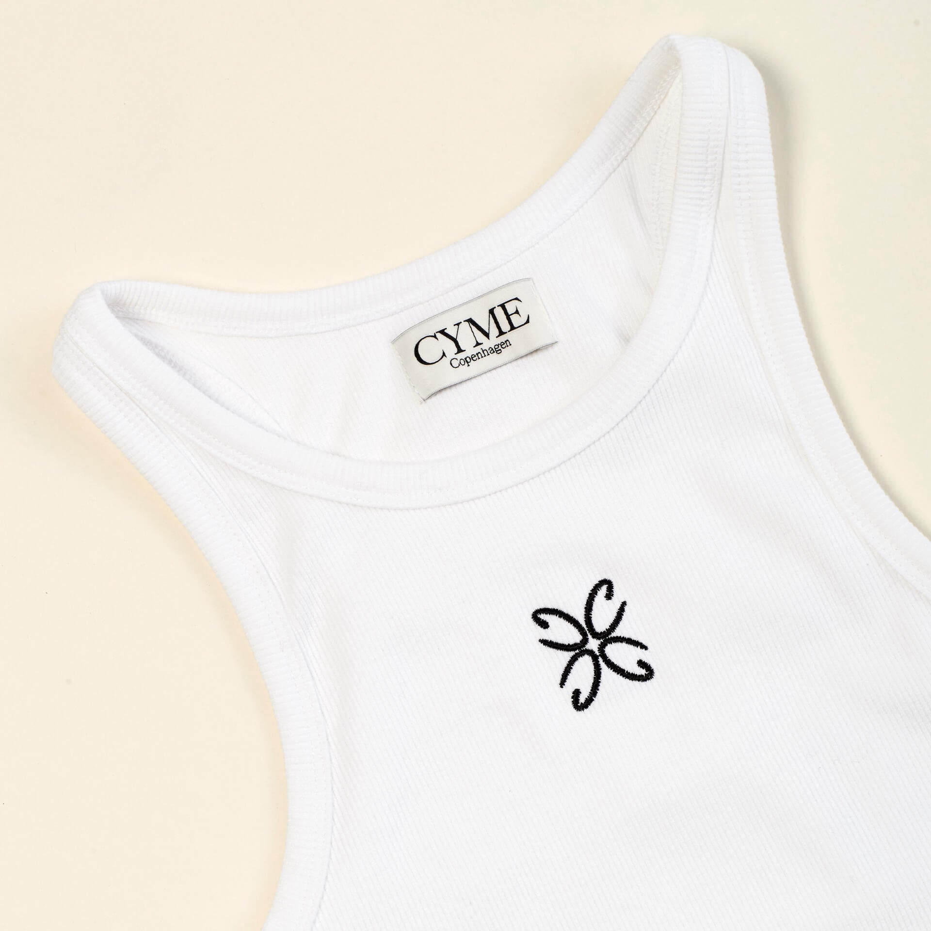 Close-up view of a white tank top with the CYME Copenhagen label sewn on the inside of the neckline and a delicate black embroidered logo on the chest. The tank top features a ribbed texture, highlighting the brand's focus on high-quality materials and timeless design.