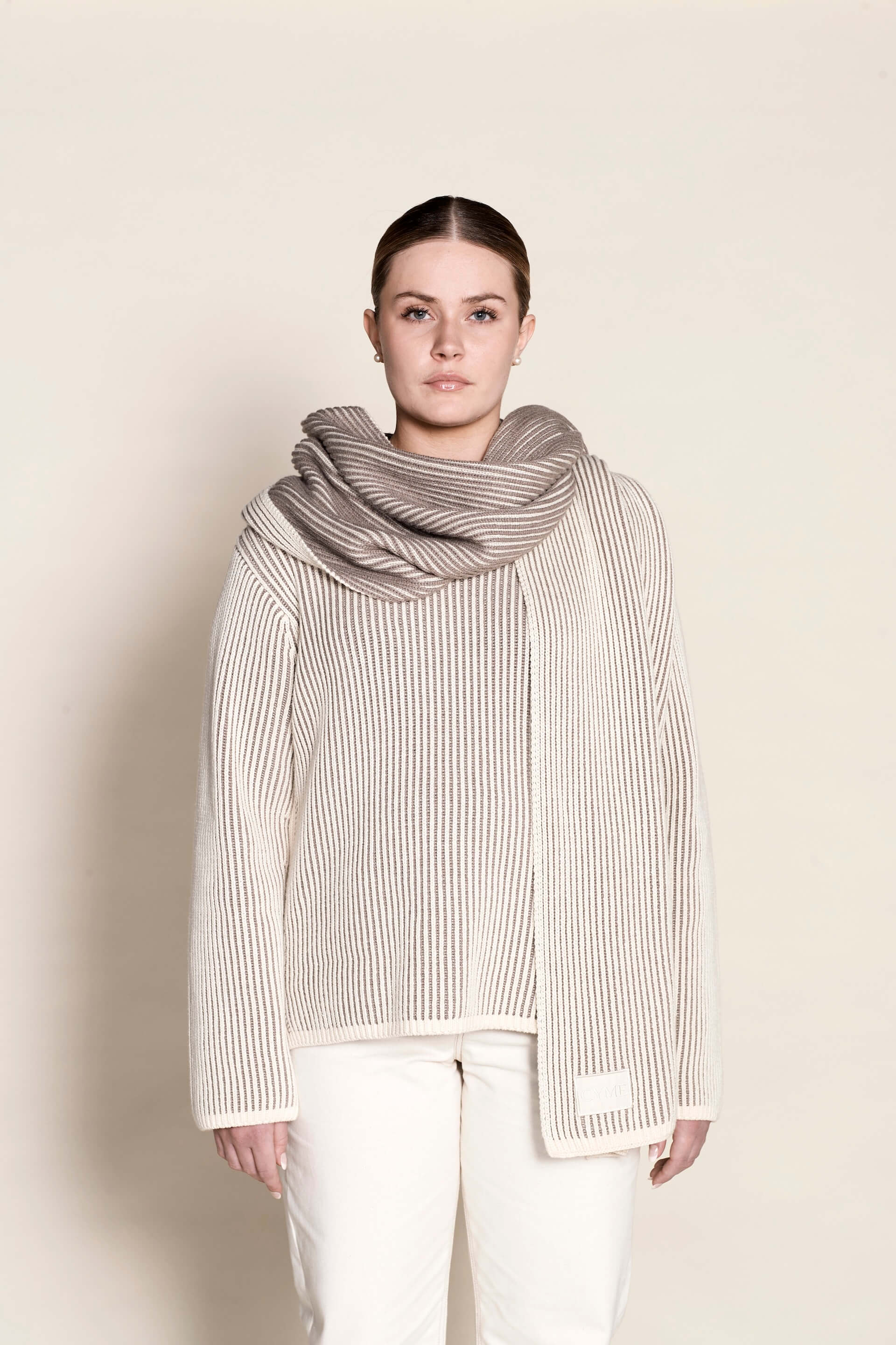 A model poses in Cyme Copenhagen's elegant cashmere knit sweater in cream, paired with a matching luxurious scarf, exemplifying the brand's commitment to sustainable fashion and natural materials.