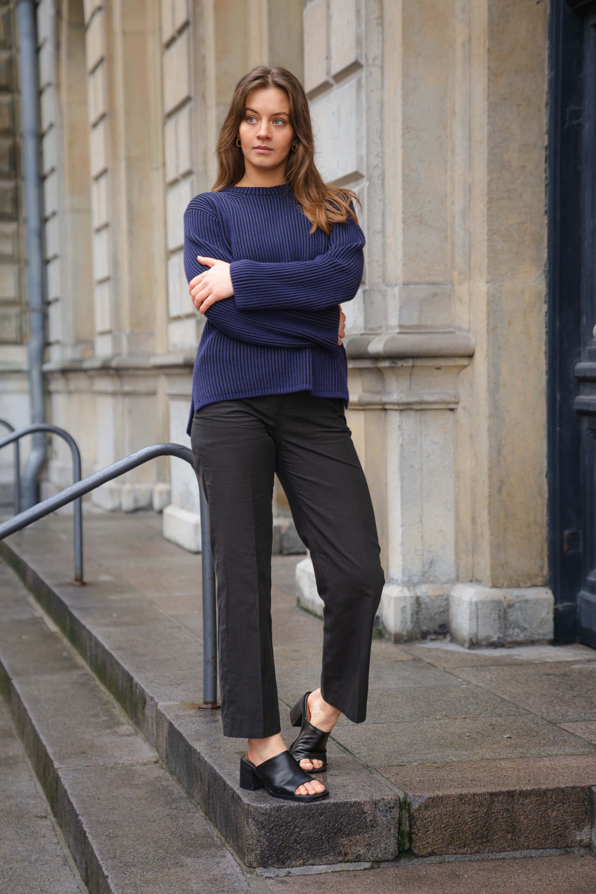 Fashion-forward look from Cyme Copenhagen, featuring a model in a deep blue ribbed jumper paired with tailored black ankle trousers and open-toe slides, reflecting the brand's dedication to crafting top fashion women's clothes with a nod to sustainable Danish design.