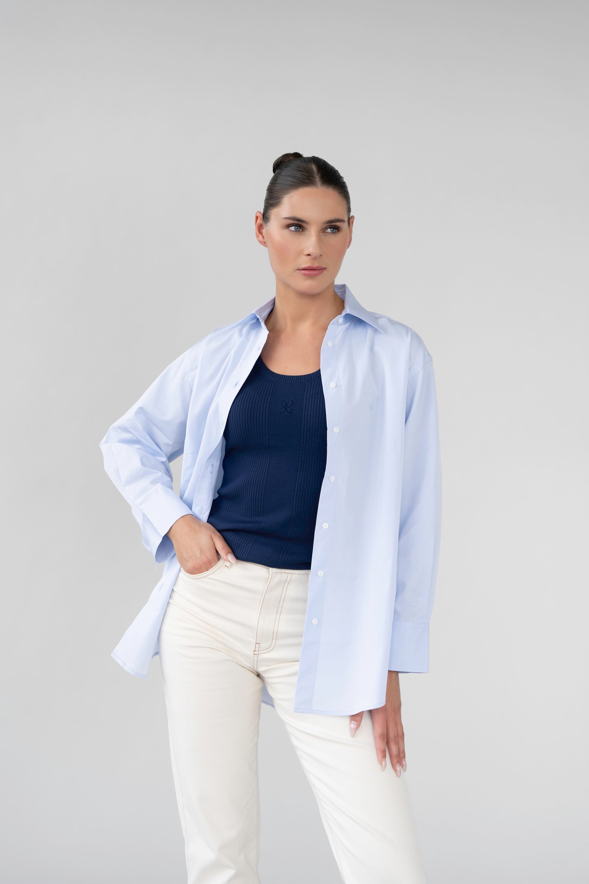 Woman wearing a light blue shirt over a navy top with white pants on a gray background