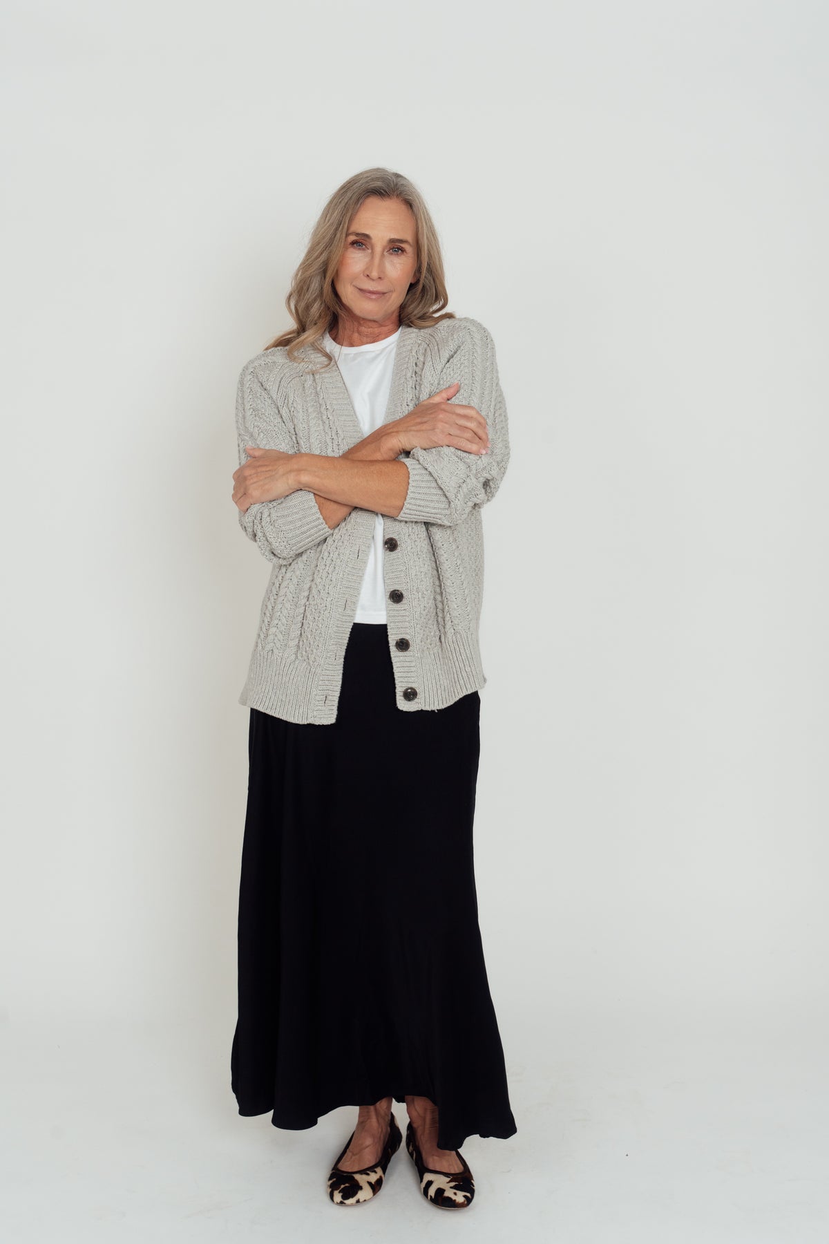 Grey Hazel Cable Knit Cardigan worn open over a white T-shirt, with sleeves pushed up and arms folded.