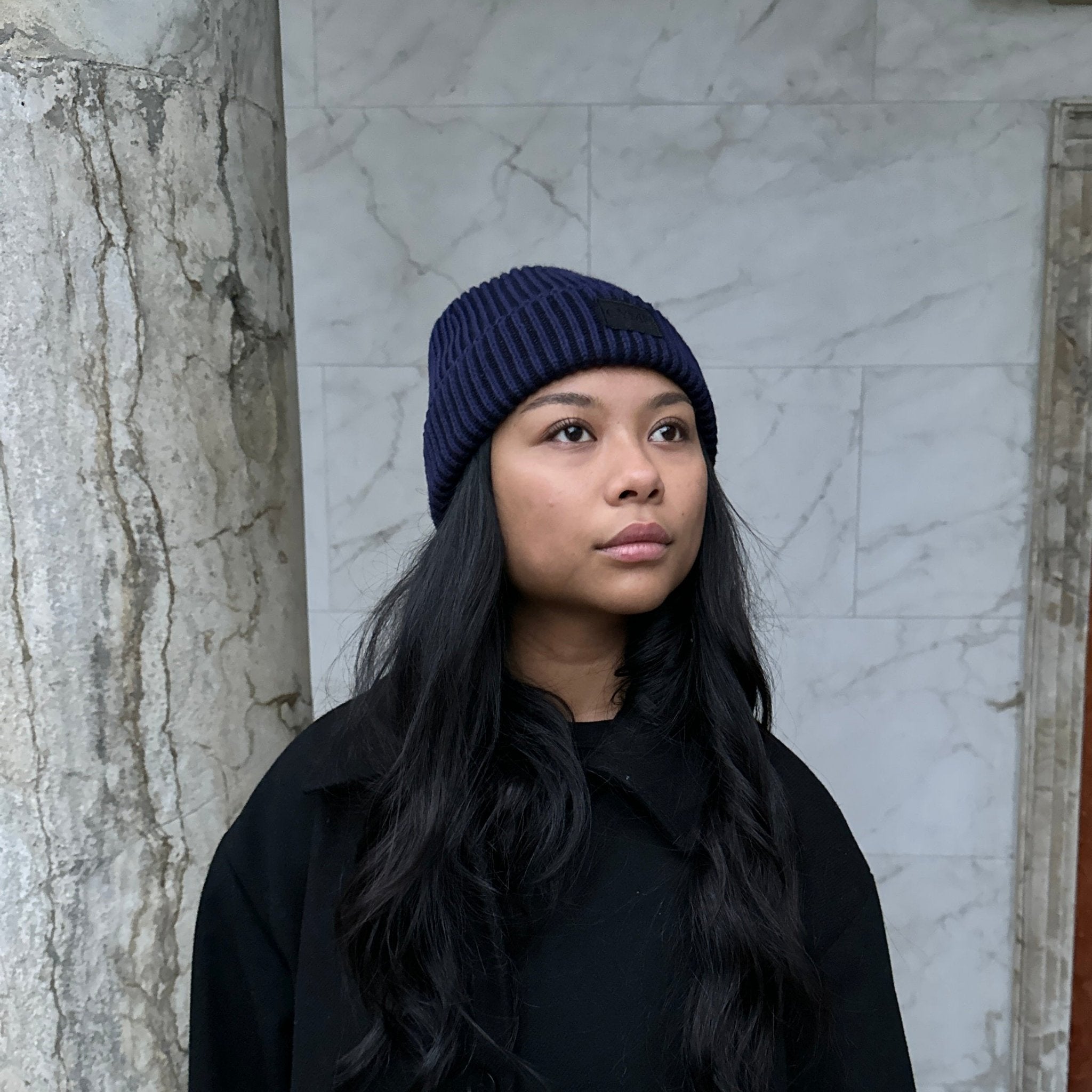 Woman wearing a dark navy knit beanie, standing in front of a marble column with long, wavy hair framing her face.