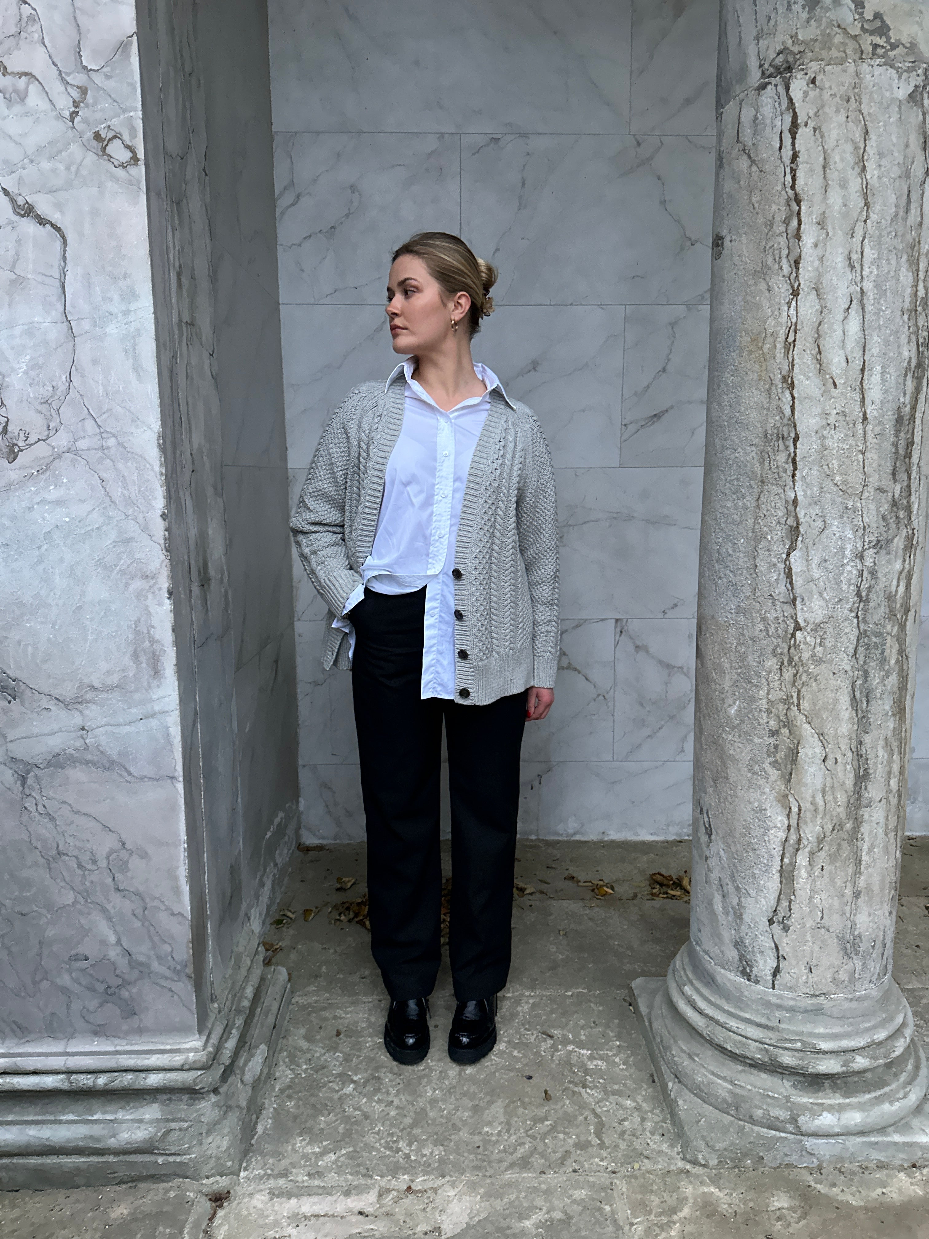 Woman wearing a gray cable-knit cardigan over a white shirt, styled with black trousers and black loafers, standing against a marble columned background.