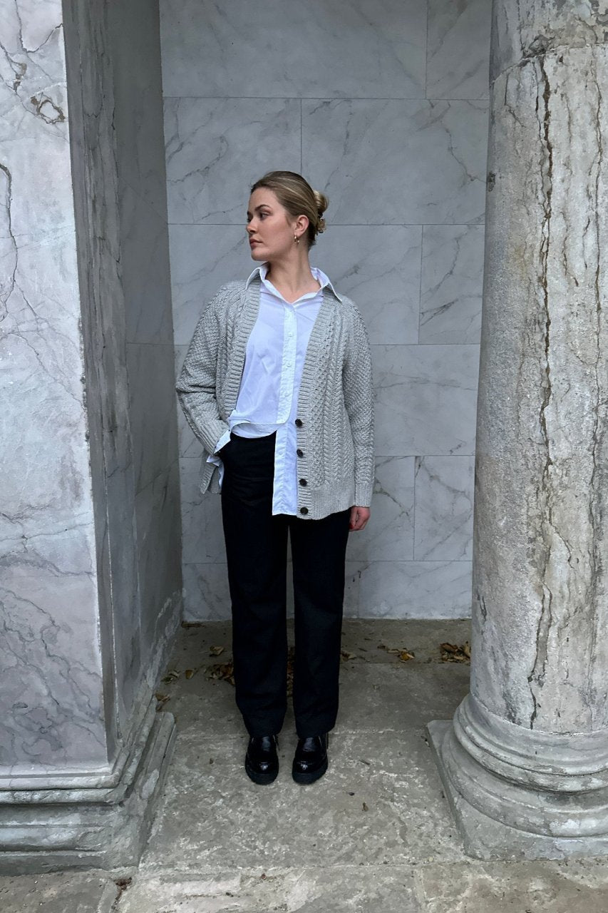 Woman wearing a gray cable-knit cardigan over a white shirt, styled with black trousers and black loafers, standing against a marble columned background.