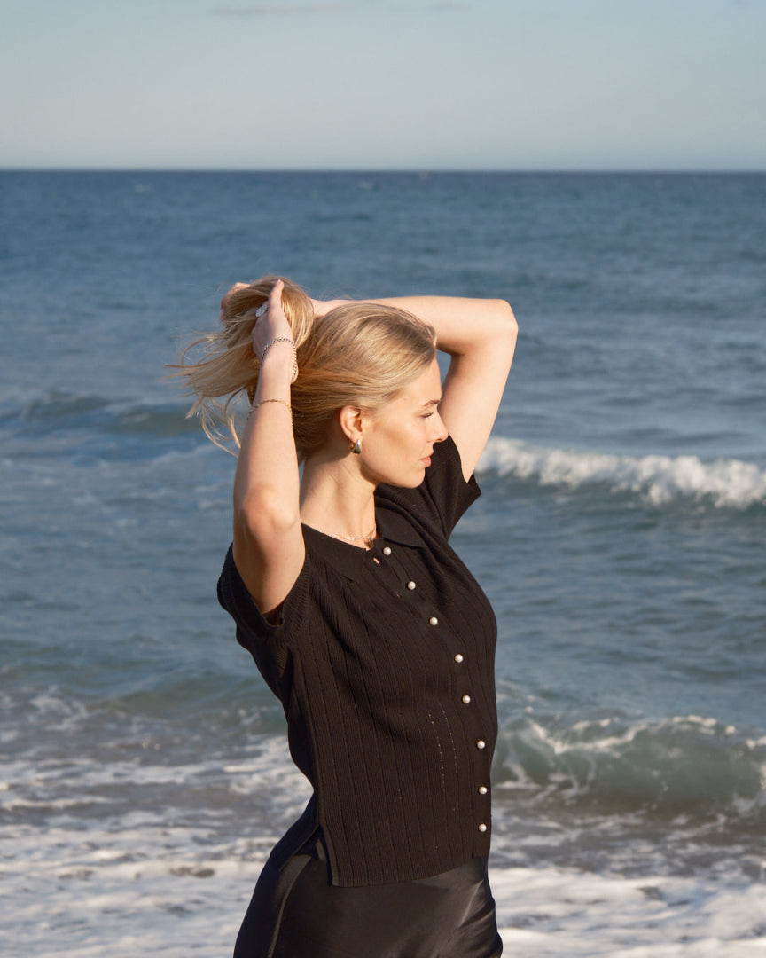 Side-profile of a blonde woman at the shoreline, arms raised to tie her hair, wearing a black rib-knit button-front top with pearl buttons.