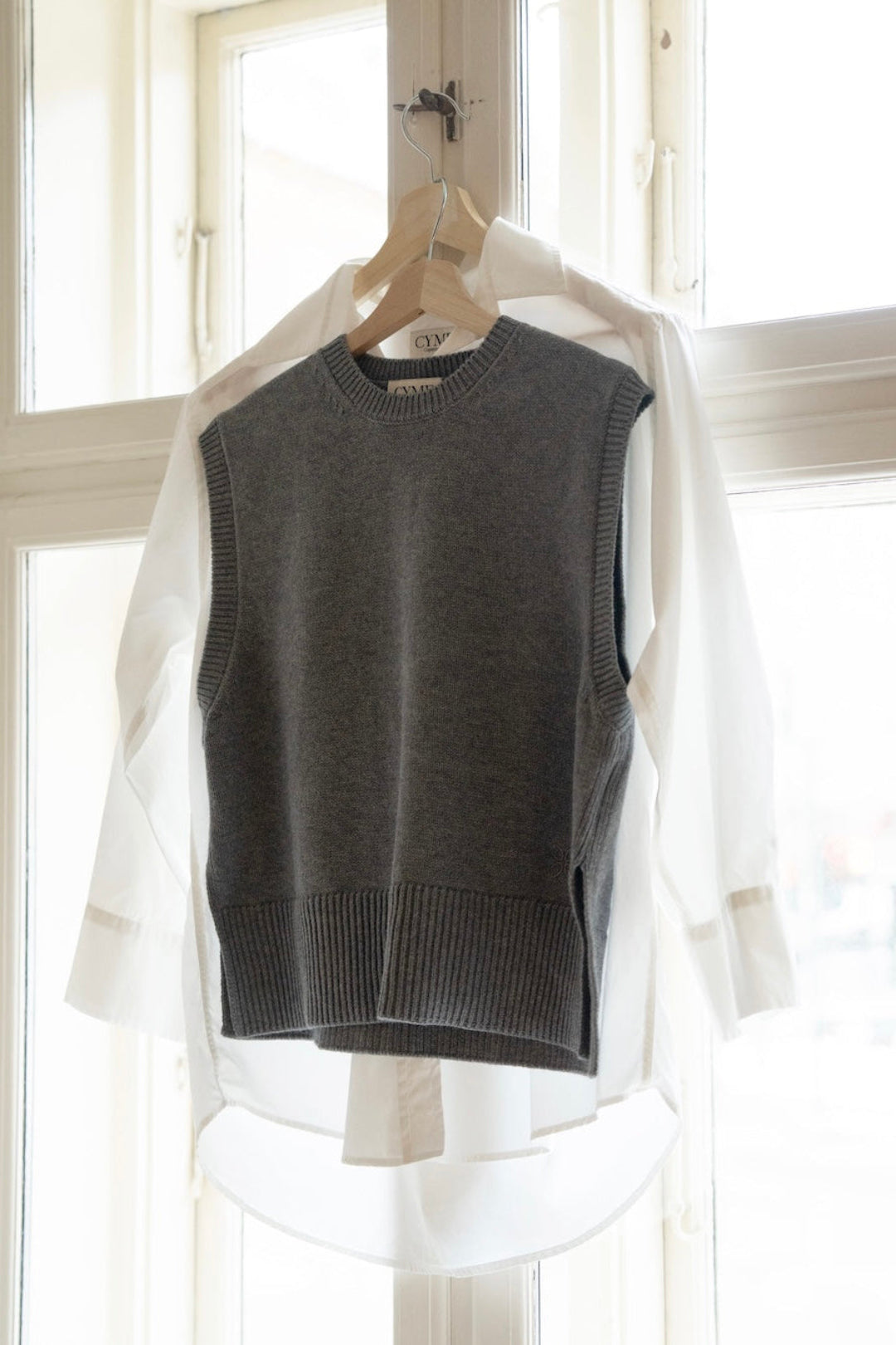 A dark gray ribbed knit sweater vest layered over a white long-sleeve shirt, hanging on a wooden hanger in front of a bright window.