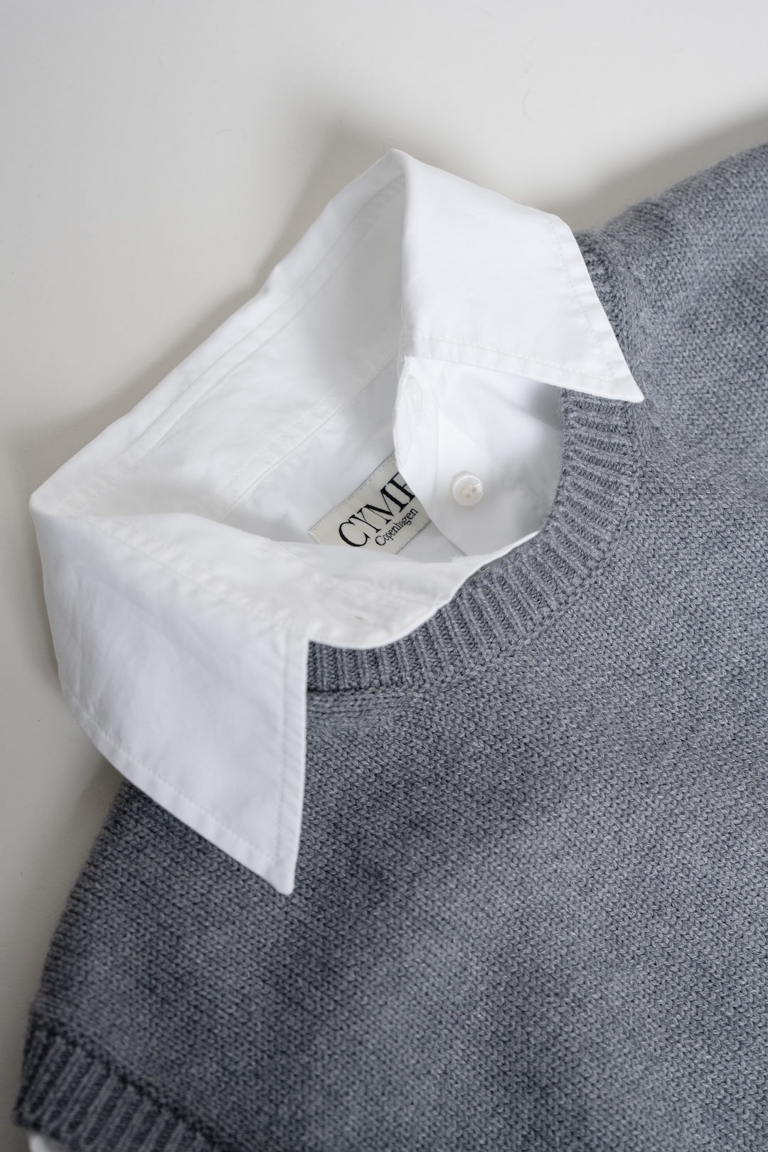 A close-up of a gray knit sweater layered over a white collared shirt, with the shirt collar folded over to show the neck label and button.