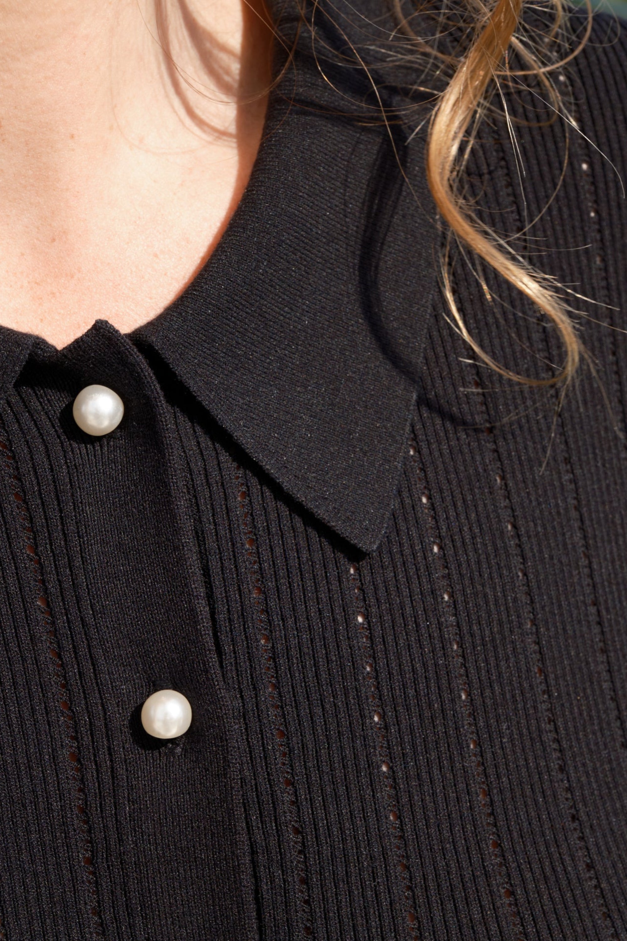 A women wearing a black polo top with pointelle holes and pearl buttons.