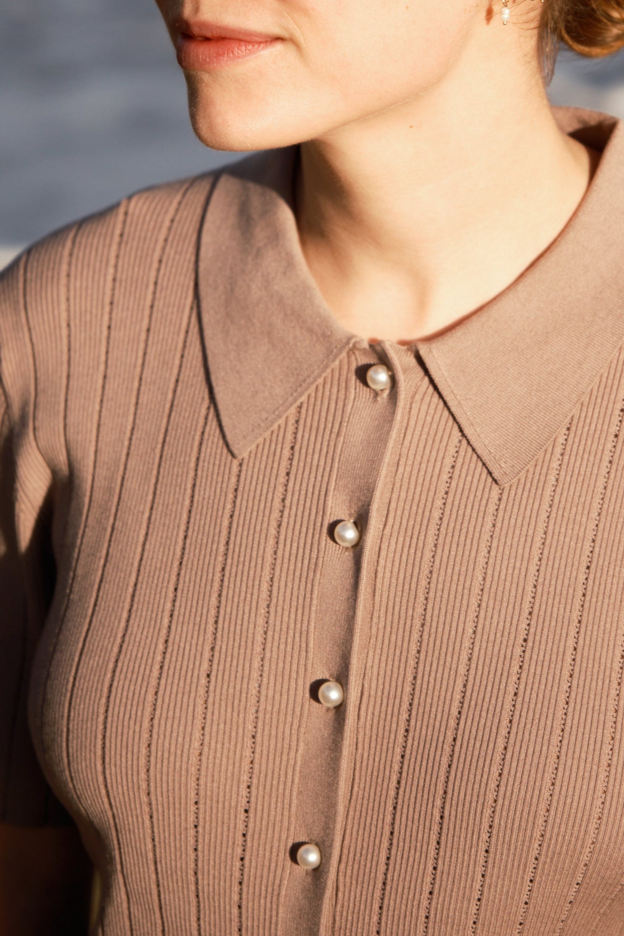 A women standing by the ocean wearing a taupe polo top with pointelle holes and pearl buttons.