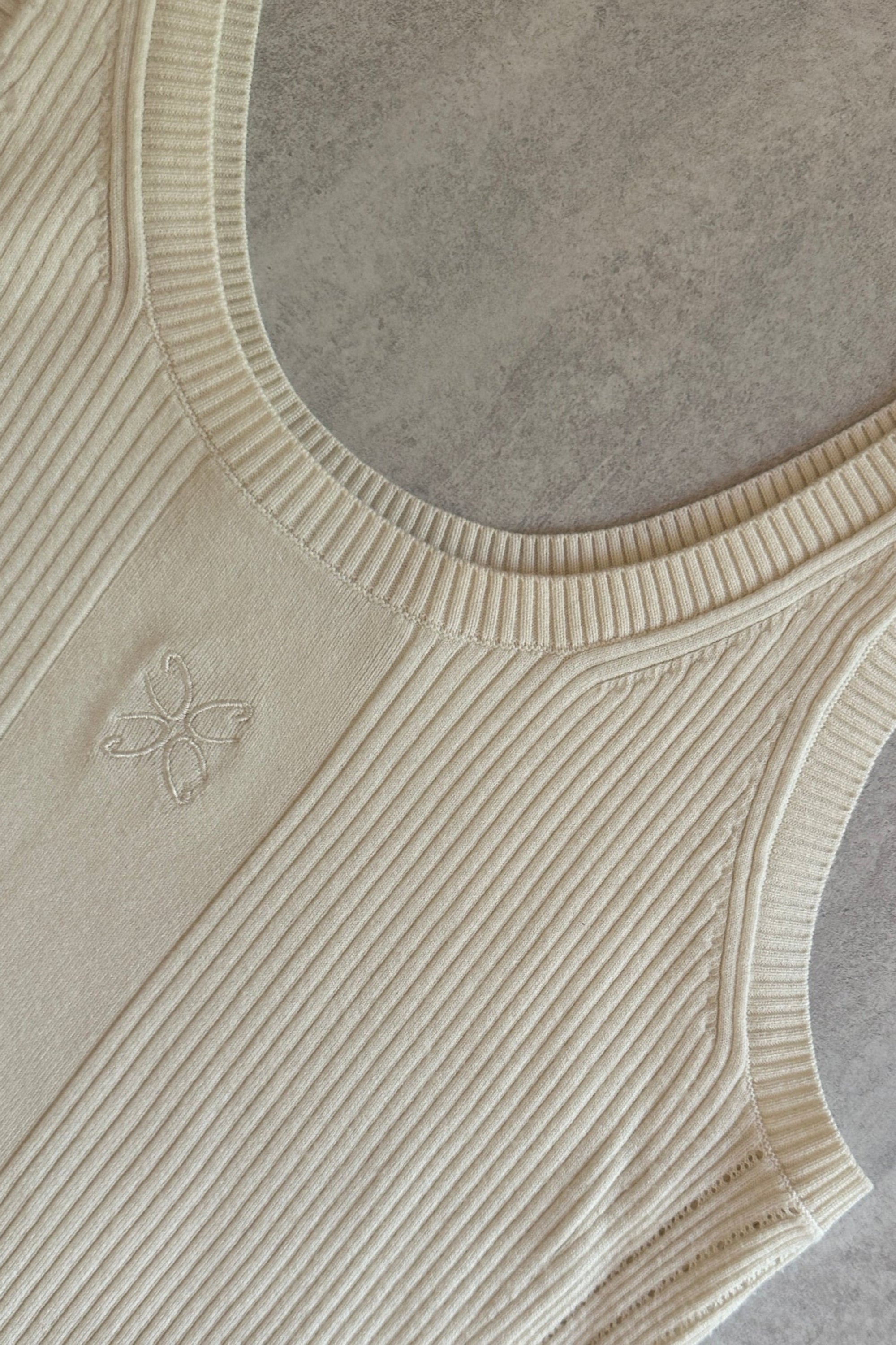Fladt produktbillede af CYME’s ribstrikkede tanktop i råhvid, med broderet blomstermotiv foran på brystet. Fremstillet i naturlige materialer og designet med en rund halsudskæring og bredde stropper.