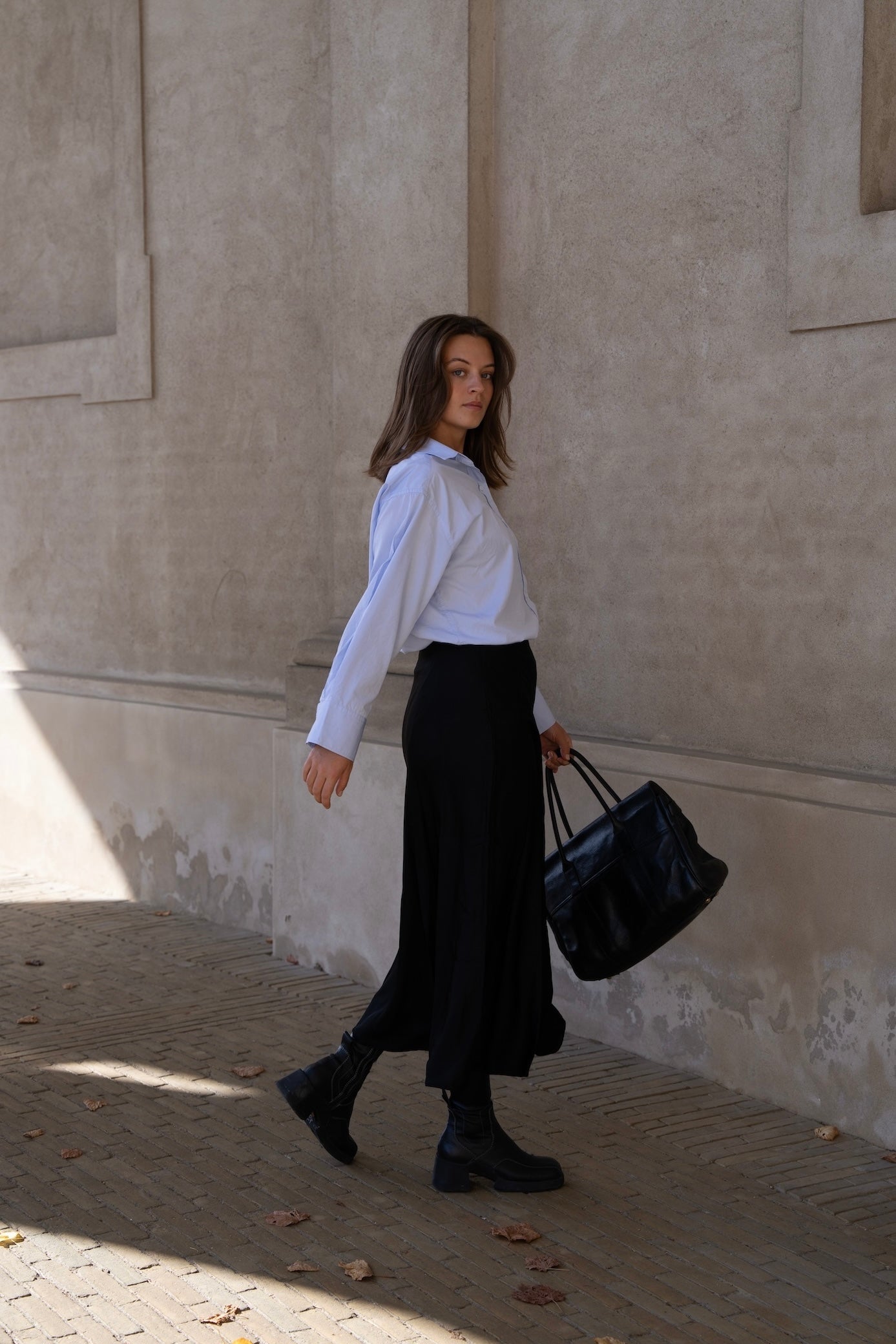 Woman in a light blue CYME Supima shirt and black satin skirt carrying a black leather bag in Copenhagen.