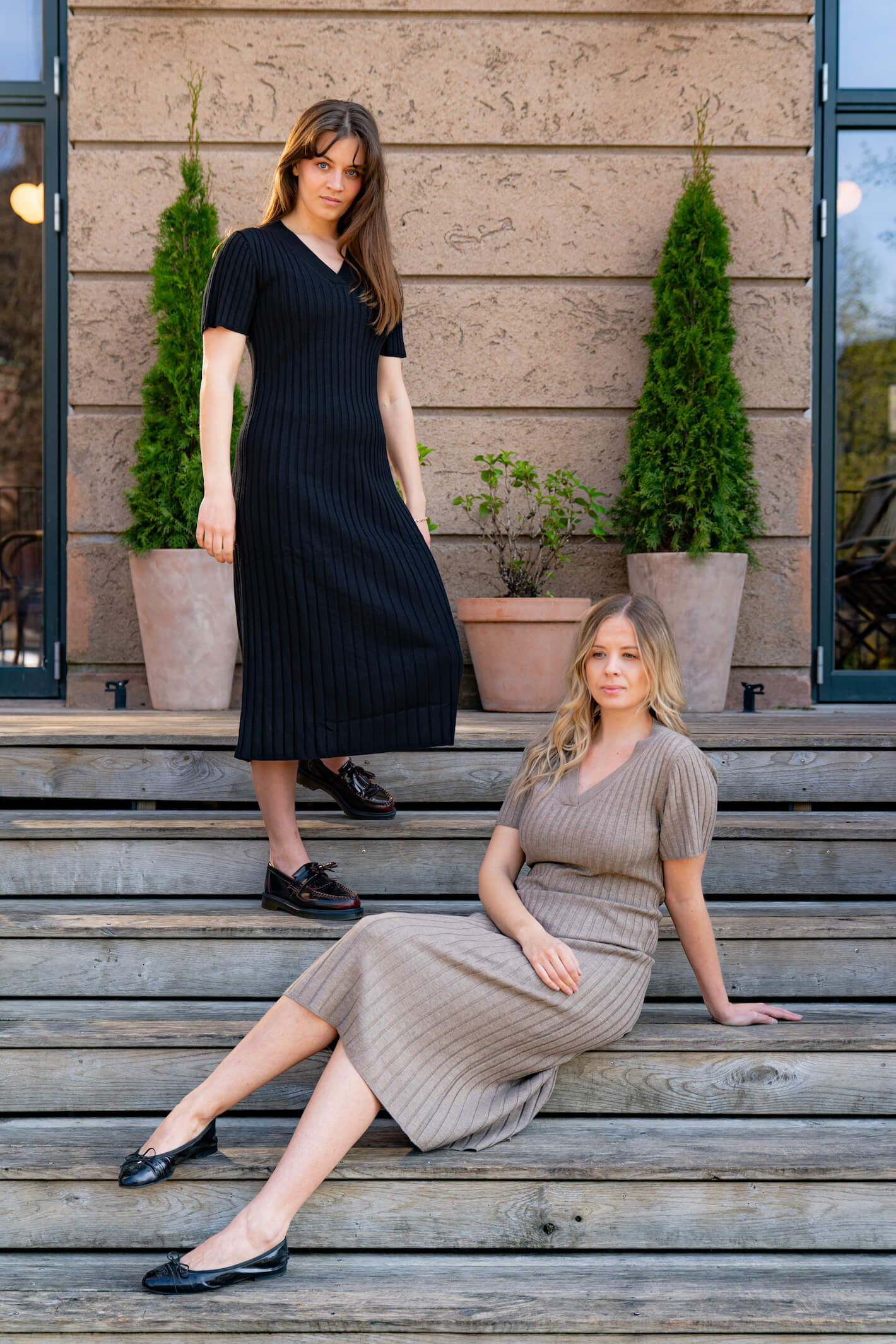 Two women modeling Cyme Copenhagen's spring/summer 2024 collection. One woman stands on wooden steps wearing a black ribbed dress and black loafers. The other woman sits on the steps wearing a beige ribbed dress and black ballet flats. The backdrop features a textured wall with potted green plants, creating a stylish and natural setting.