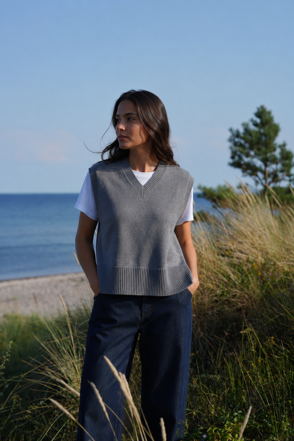 A woman standing outdoors by the beach wearing a gray sleeveless knit sweater vest over a white t-shirt and dark blue wide-leg pants.