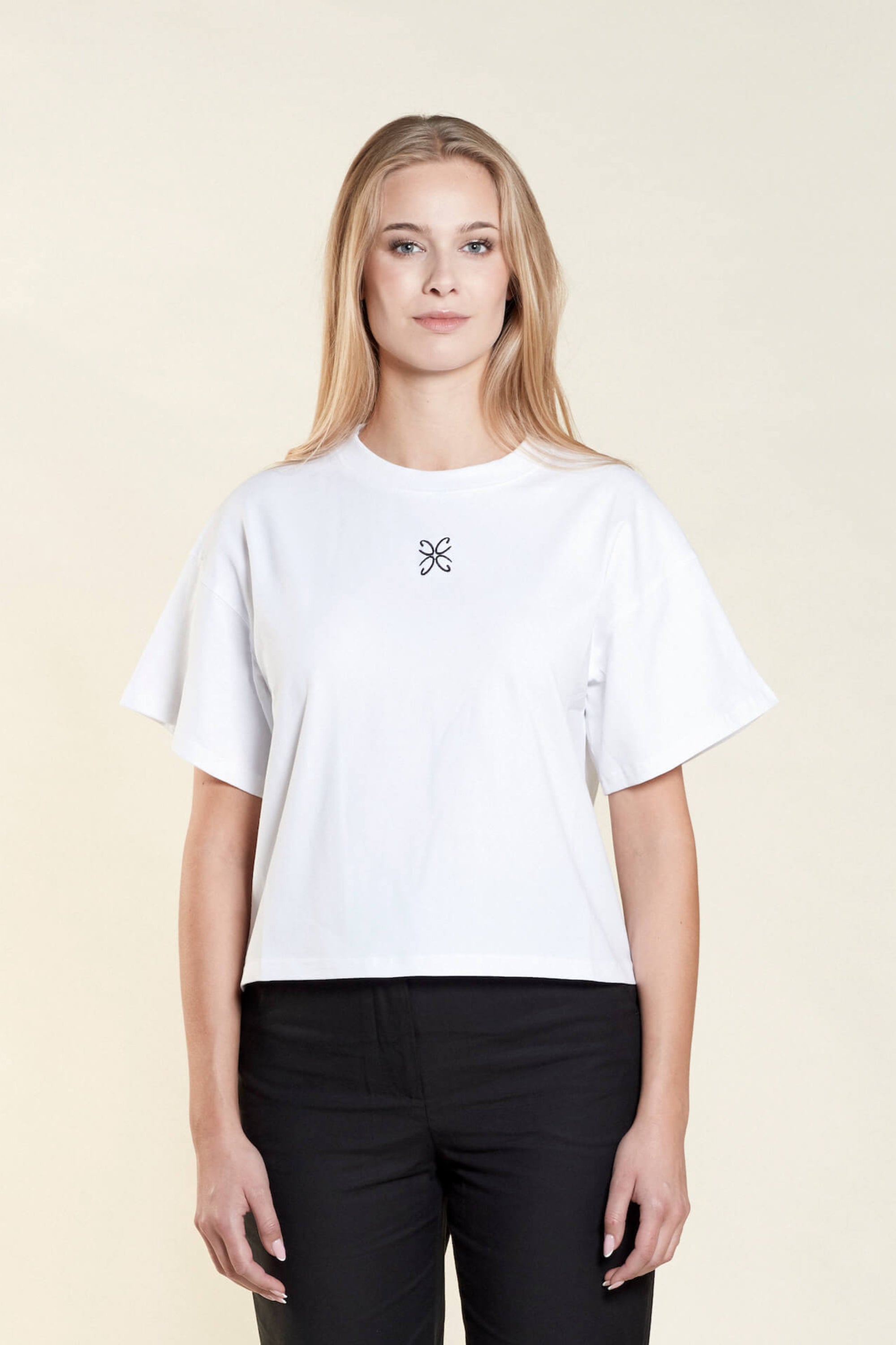 Woman wearing a white t-shirt from Cyme Copenhagen's spring/summer collection. The t-shirt features a simple design with a black logo in the center. The model stands against a neutral background, highlighting the minimalist and stylish appeal of the garment.