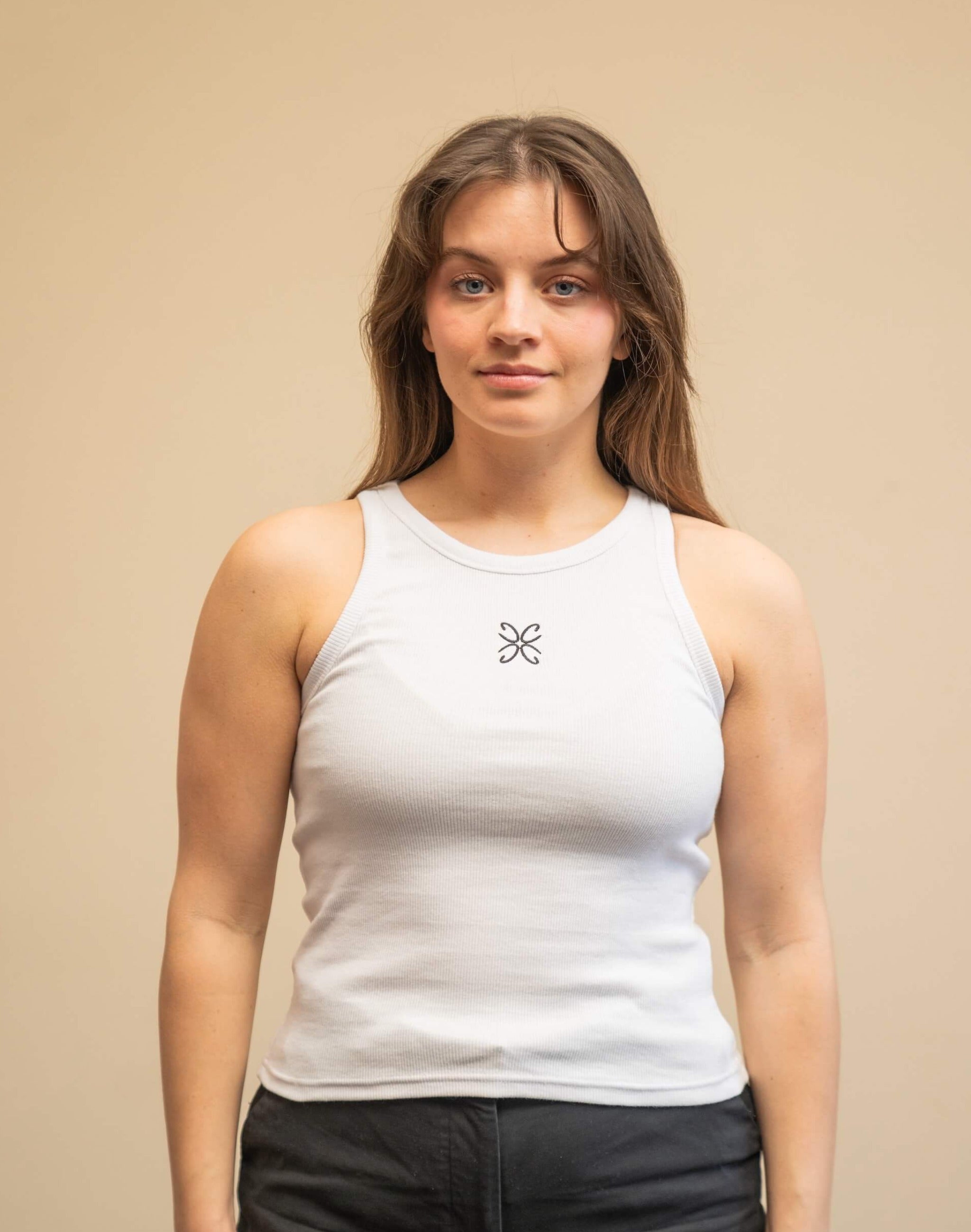 Woman wearing a white tank top from Cyme Copenhagen's spring/summer collection. The tank top features a simple design with a black logo in the center. The model stands against a neutral background, emphasizing the minimalist and stylish appeal of the garment.