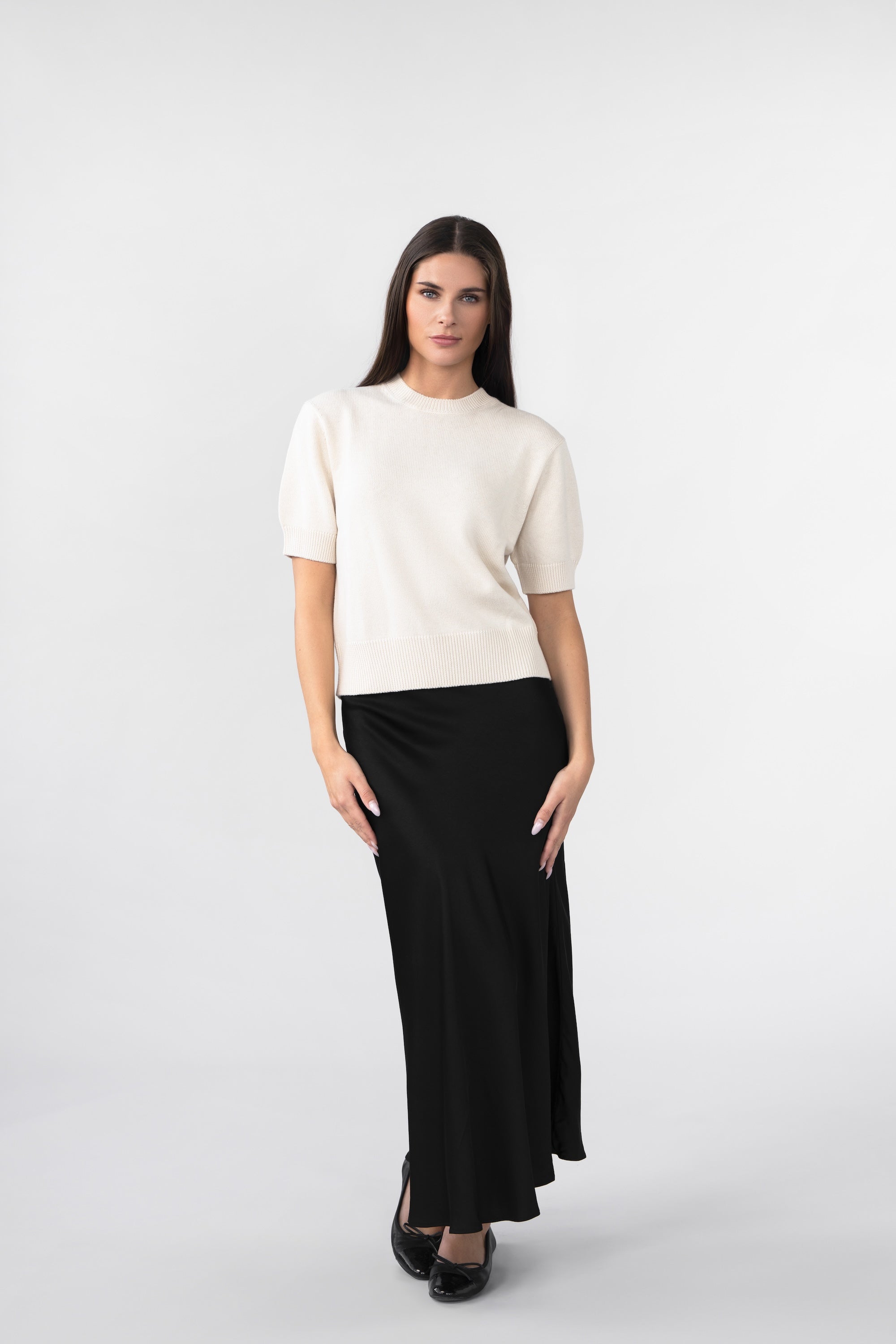 A woman wearin a white knitted tee and a black satin skirt against a gray background.