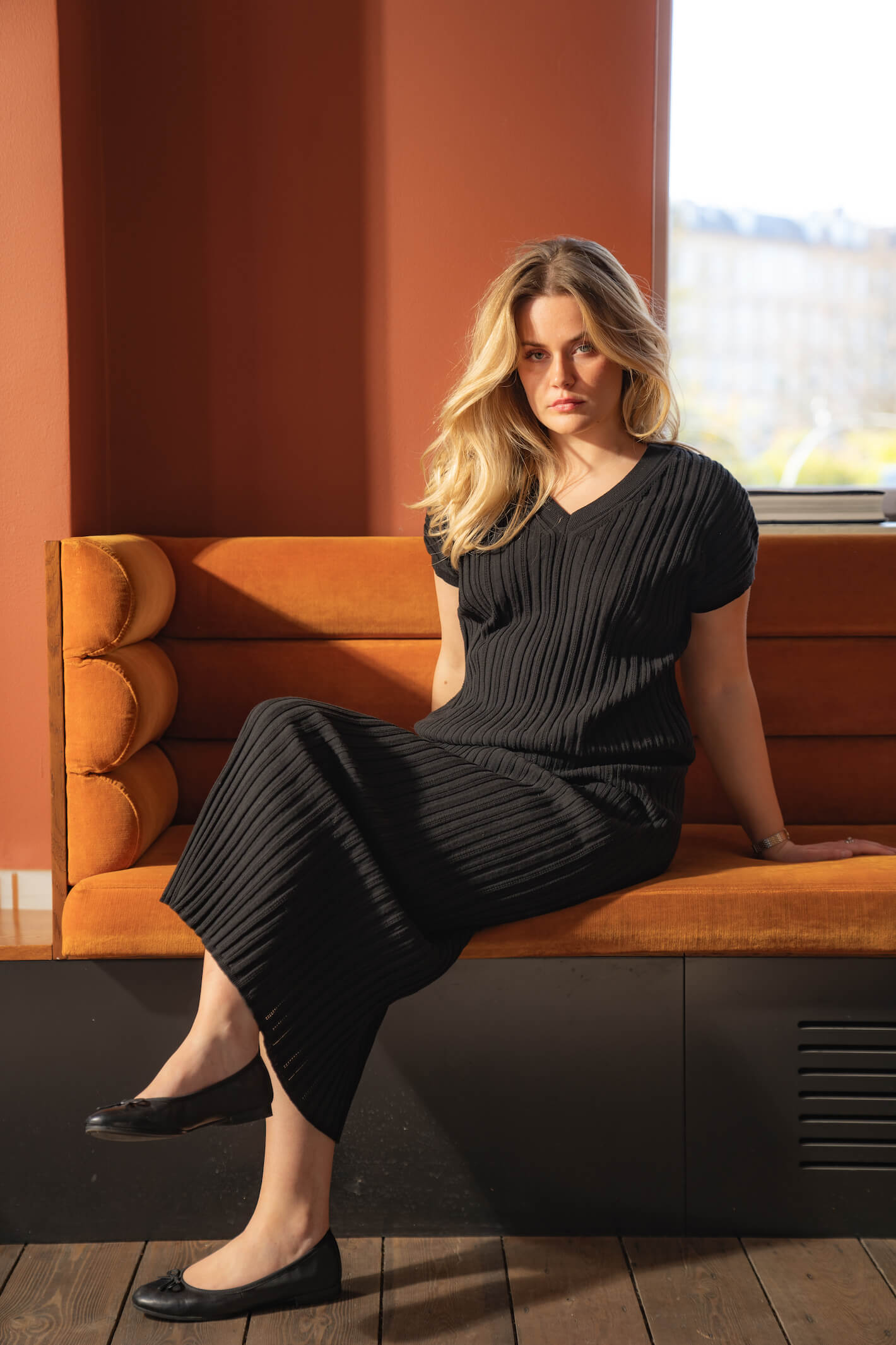 Woman sitting on an orange couch wearing a black ribbed dress by Cyme Copenhagen. She has blonde hair and black ballet flats, with an orange and sunlit background. The scene highlights the elegance and comfort of the Cyme Copenhagen spring/summer 2024 collection.
