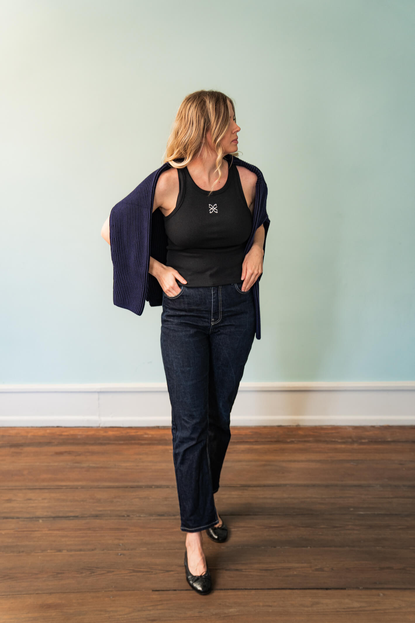 Woman modeling Cyme Copenhagen's spring/summer 2024 collection. She wears a black tank top with a white logo, dark denim jeans, and black ballet flats. A dark blue cardigan is draped over her shoulders. The setting features a light teal wall and wooden floor, highlighting the casual yet stylish look of the collection.