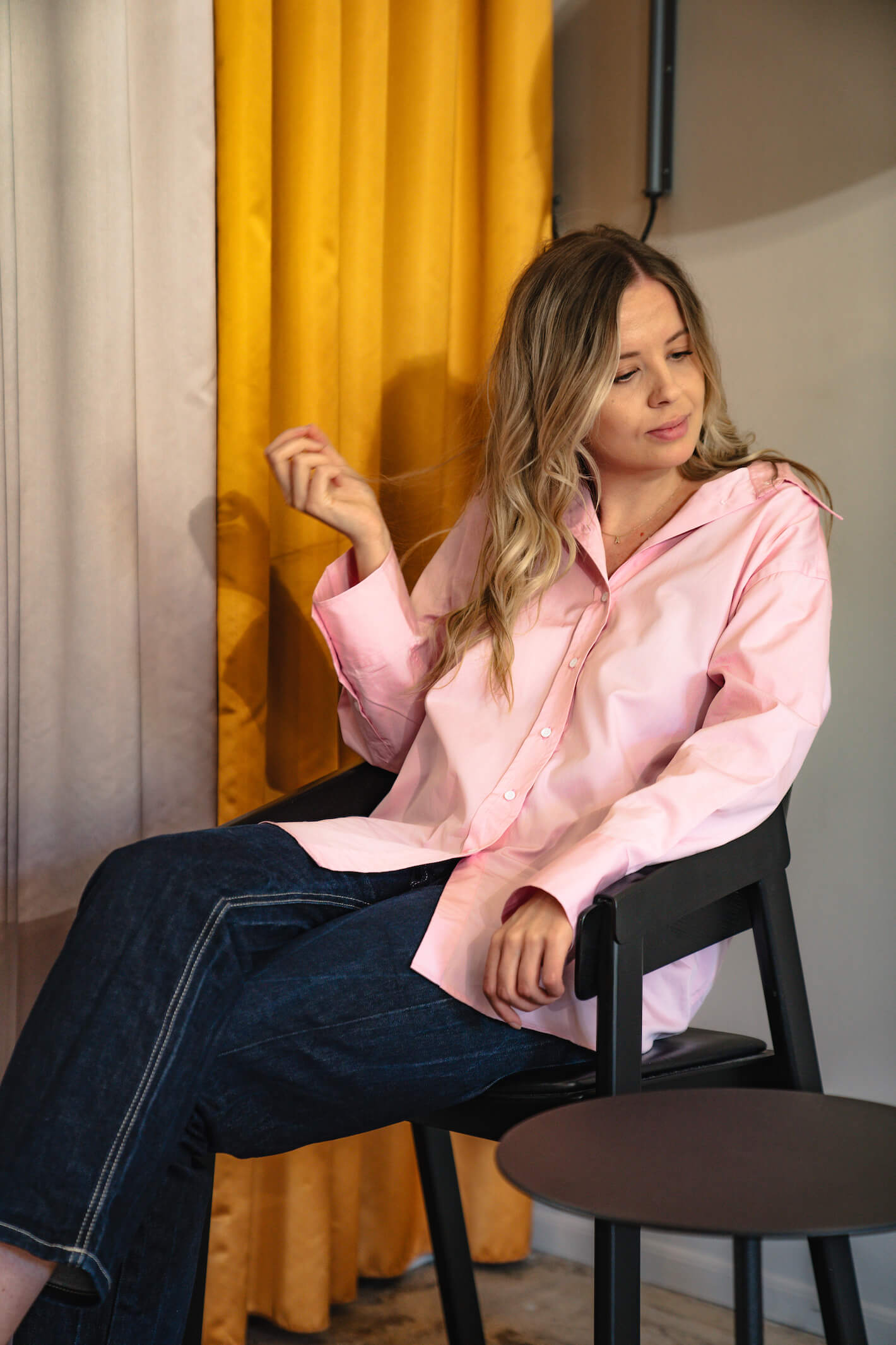 Woman sitting on a black chair wearing a pink button-down shirt and dark denim jeans from Cyme Copenhagen's spring/summer 2024 collection. She has blonde hair and the setting features a background with yellow and white curtains, adding a touch of warmth and elegance to the stylish, casual look.