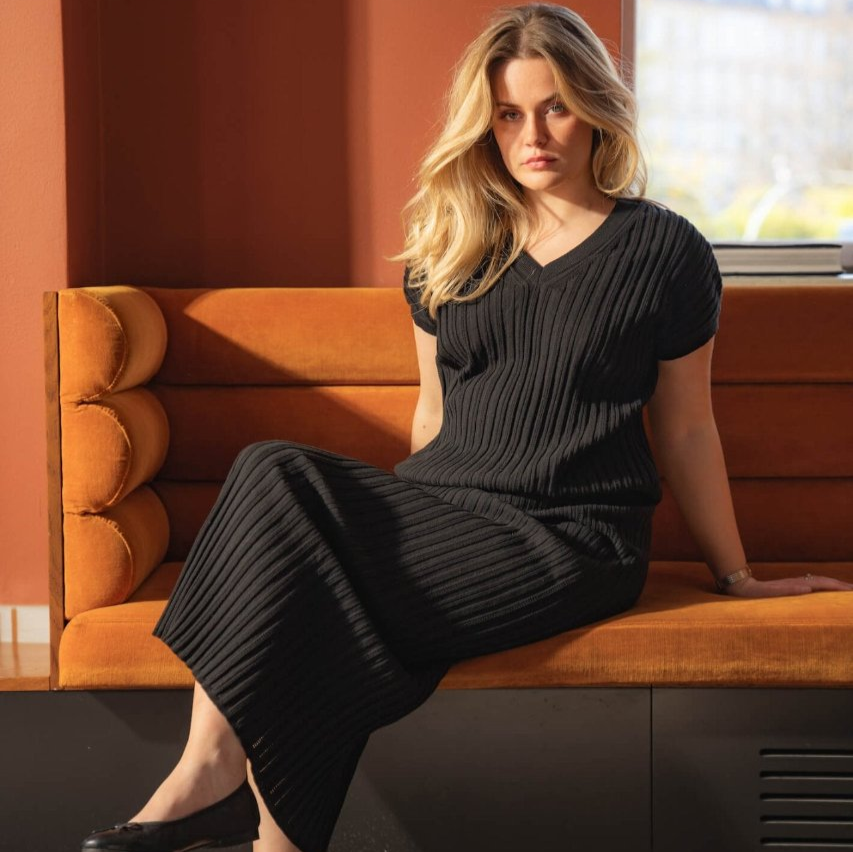 Woman sitting on an orange couch wearing a black ribbed dress by Cyme Copenhagen. She has blonde hair and black ballet flats, with an orange and sunlit background. The scene highlights the elegance and comfort of the Cyme Copenhagen spring/summer 2024 collection.